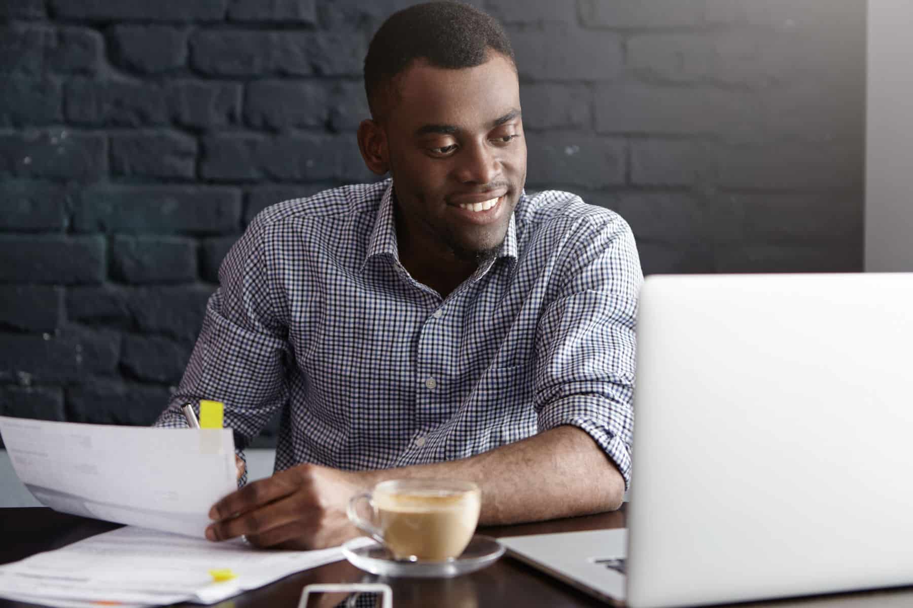 Happy confident young African-American businessman in formal wear filling in papers while managing finances at cafe during lunch break, sitting at table in front of open laptop, having cappuccino